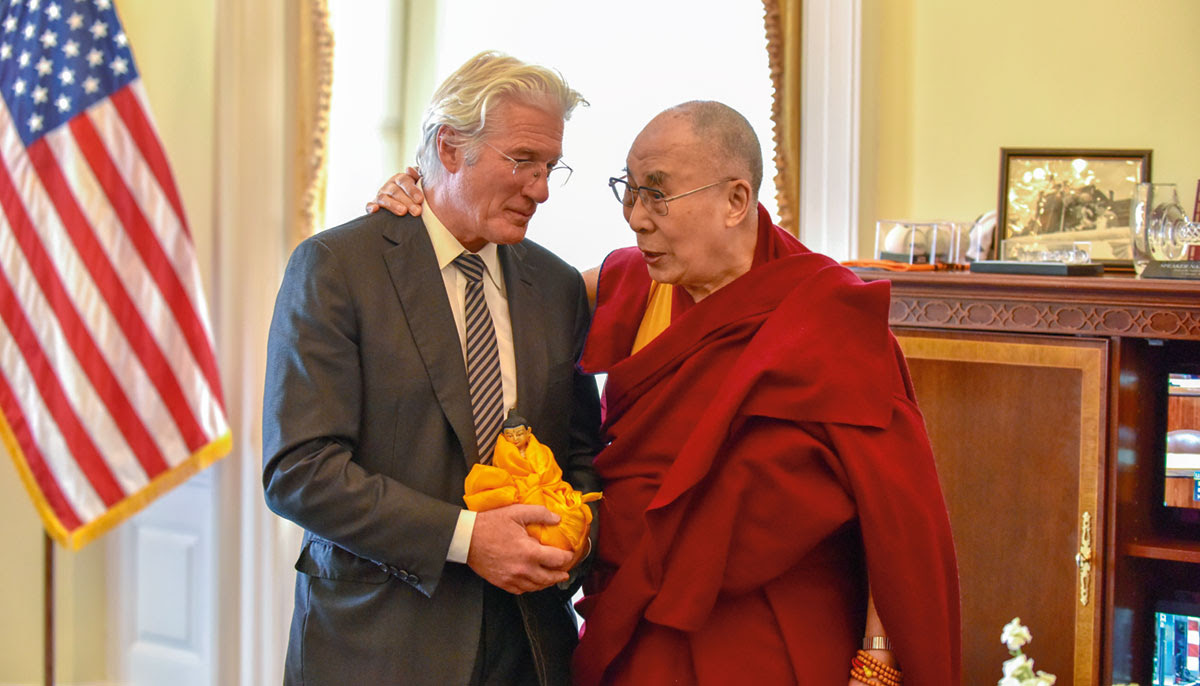 A man dressed in a suit stands near the Dalai Lama, wearing red monk's robes.