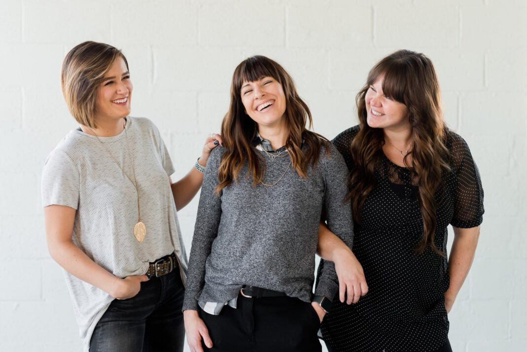 3 femmes souriantes et debout à côté d'un mur blanc